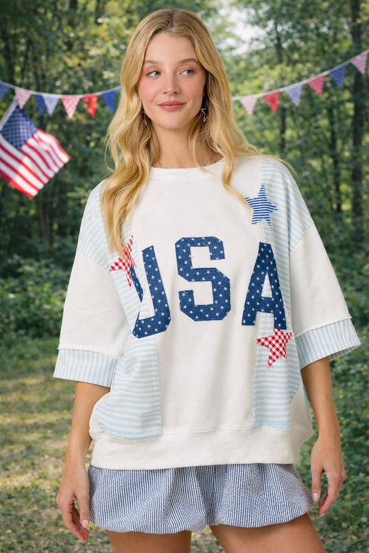 Woman wearing a white sweater with 'USA' printed on it, standing outdoors with American flags in the background.