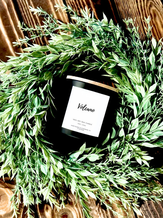 Black candle labeled 'Velano' surrounded by green foliage on a wooden surface