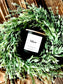 Black candle labeled 'Velano' surrounded by green foliage on a wooden surface