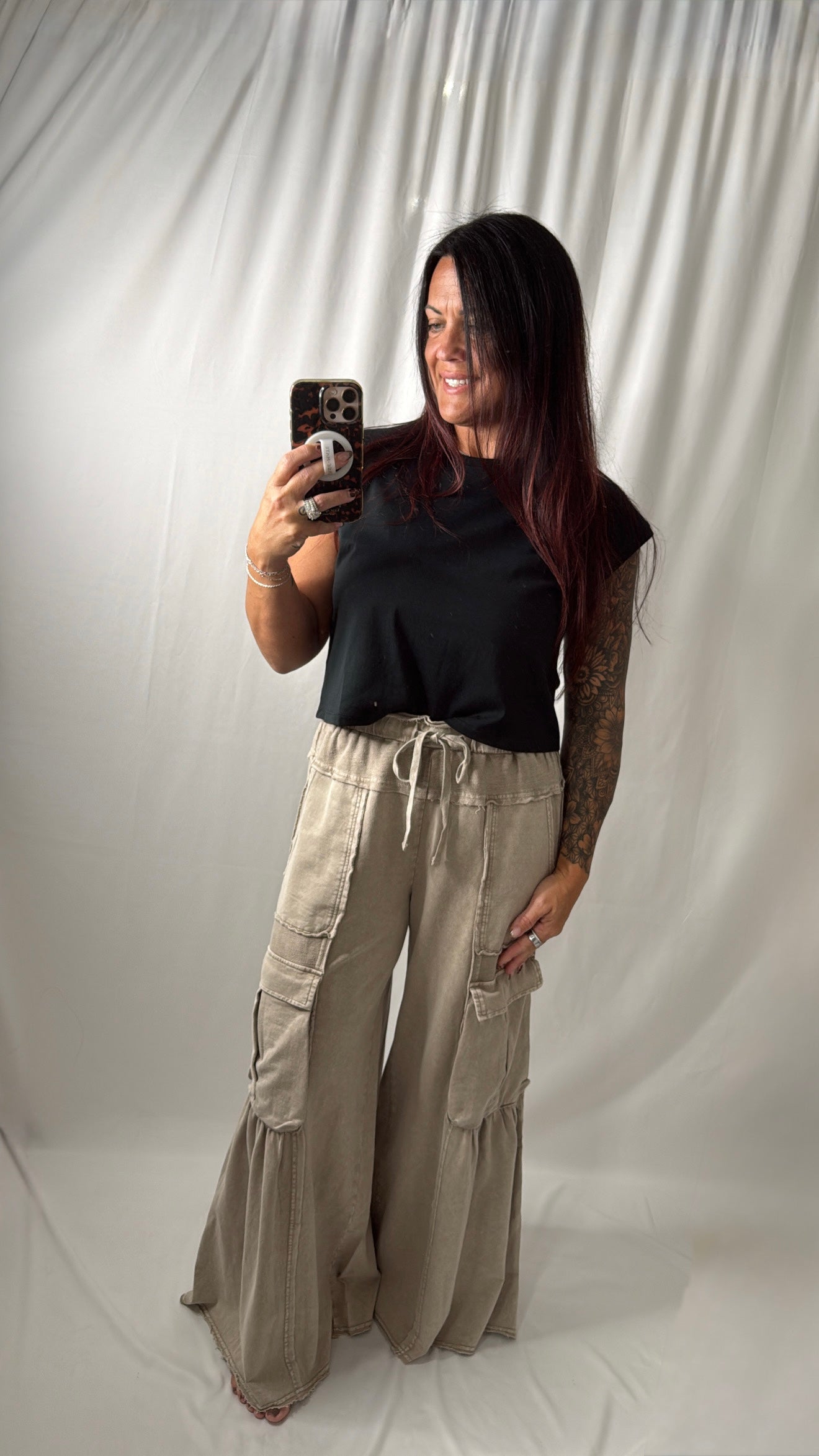 A woman takes a mirror selfie wearing a black short-sleeve top and beige wide-leg cargo pants with a drawstring waist, set against a white draped fabric backdrop.