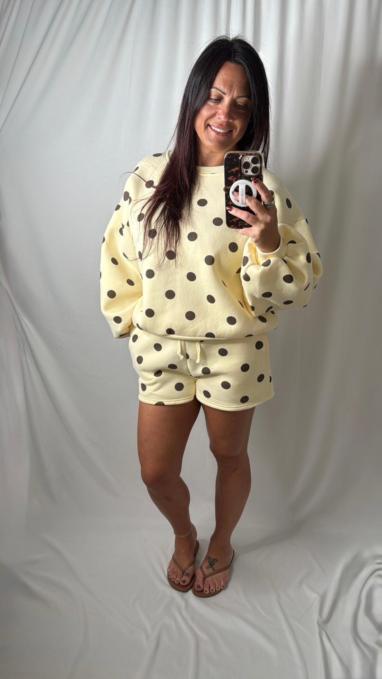 A woman takes a mirror selfie wearing a butter yellow-colored two-piece loungewear set with brown polka dots, consisting of a sweatshirt and matching shorts, against a draped white fabric backdrop.
