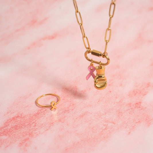 Gold necklace with a pink ribbon charm and matching ring on a pink marble background
