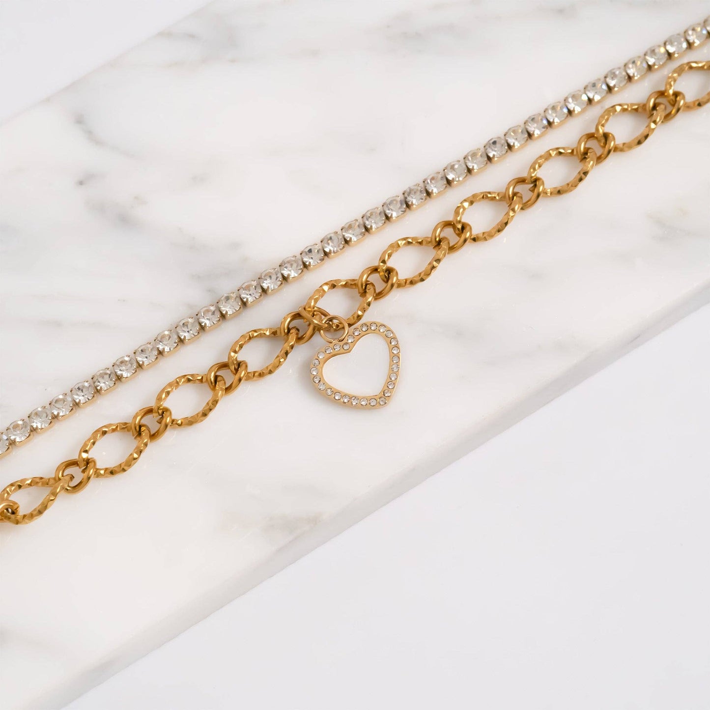 Two gold necklaces with heart charm and clear gemstones on a marble surface