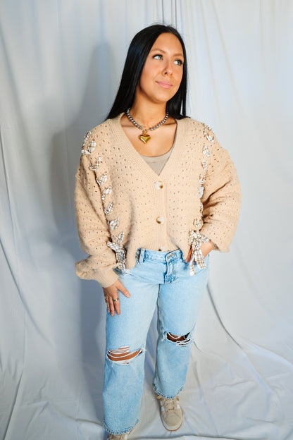 Woman wearing a beige cardigan with decorative elements, light blue jeans, and beige shoes against a plain background.