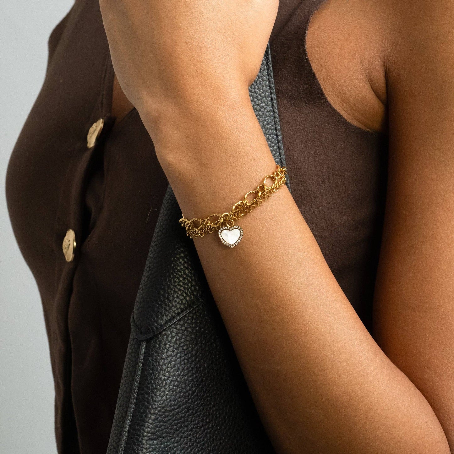 Gold bracelet with a heart charm on a person's wrist, wearing a brown jacket.