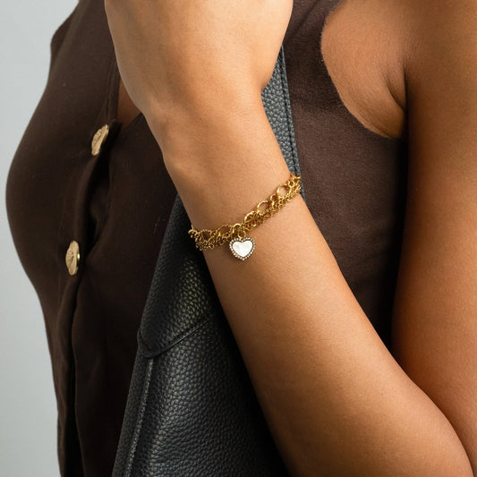Gold bracelet with a heart charm on a person's wrist, wearing a brown jacket.