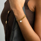 Gold bracelet with a heart charm on a person's wrist, wearing a brown jacket.