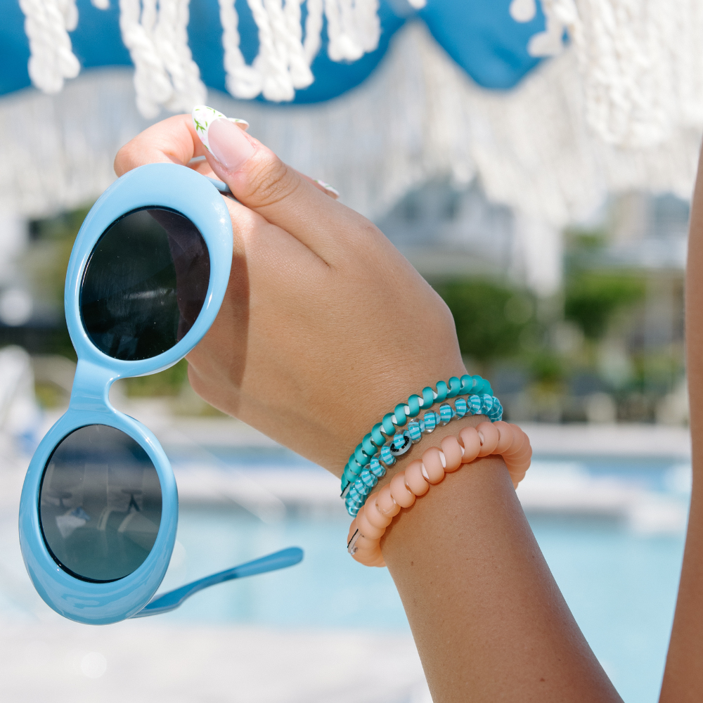 Hand holding blue sunglasses with a blurred pool and deck chair in the background with 3 teleties hair slips on the wrist