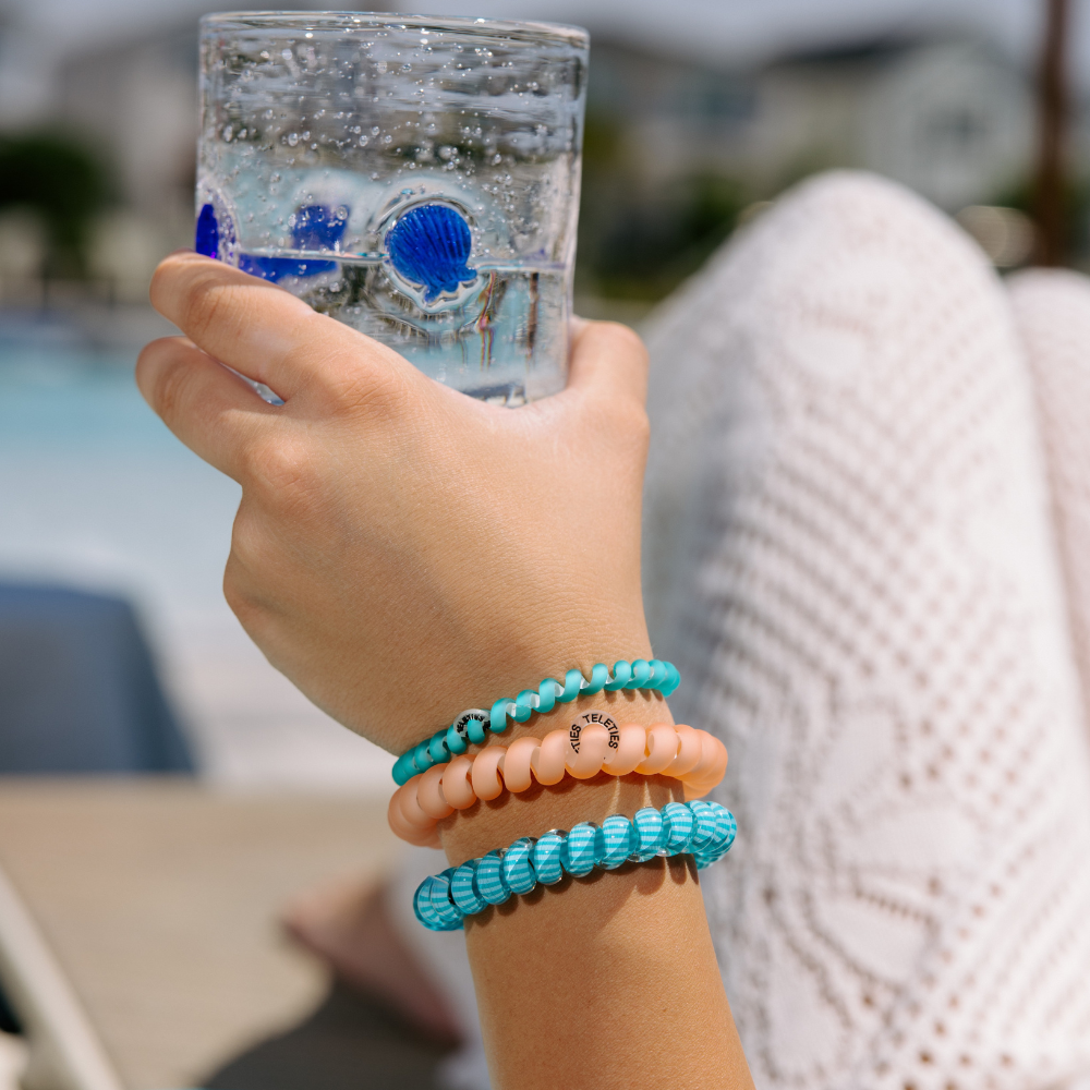 Hand holding a glass of water with a blurred outdoor background with 3 teleties hair ties on her hand